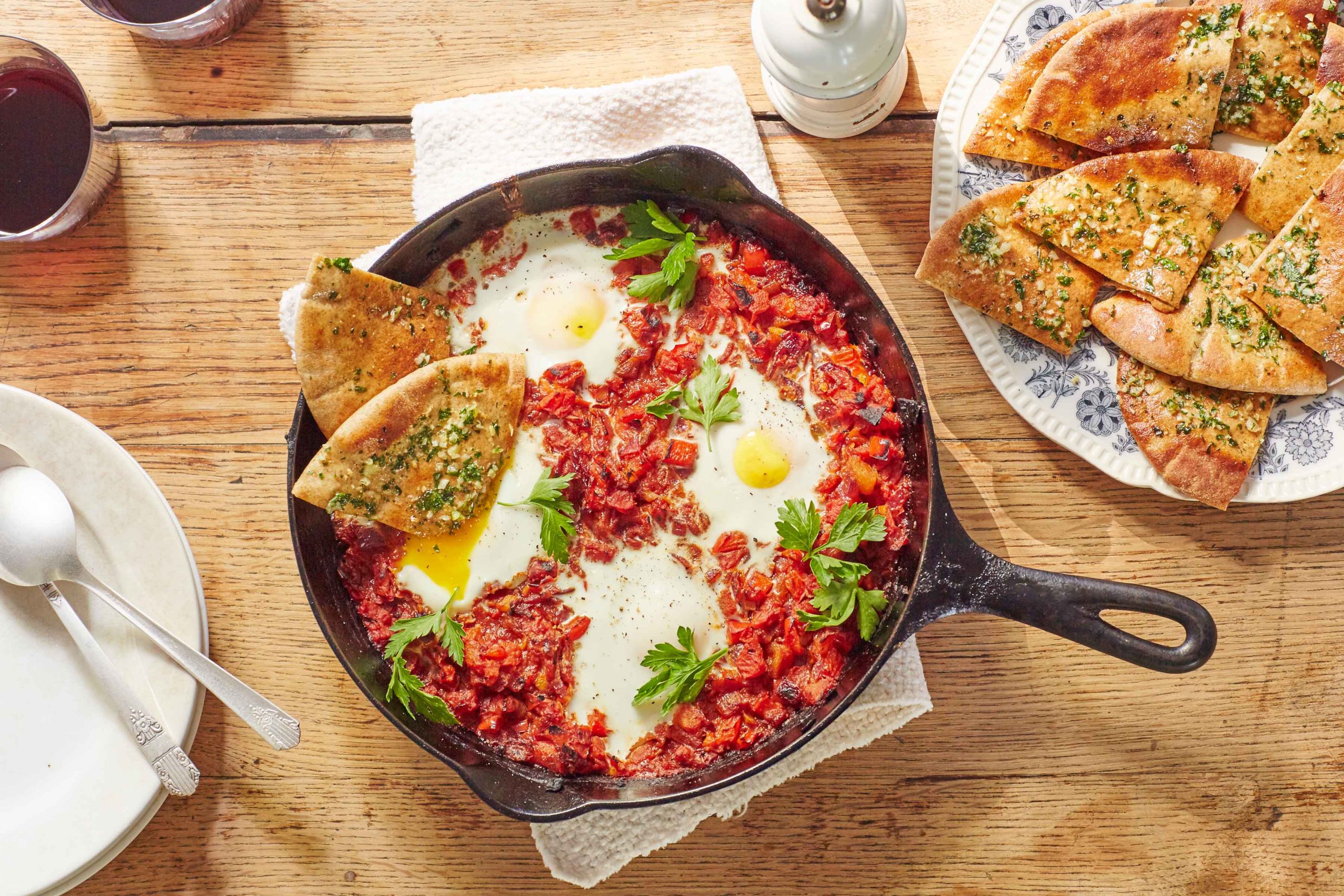 Shakshuka, a Middle Eastern tomato and egg dish, baked in a cast iron skillet with a plate of toasted pita cut in triangles and topped with herbs on the side